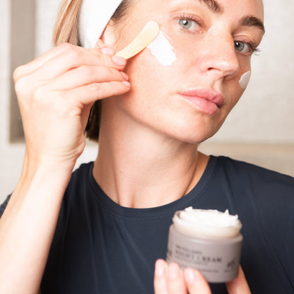 A woman applies Face Junkie Pro Collagen Night Cream to her cheek using a small wooden spatula. She wears a white headband to keep her hair back, allowing full focus on her skincare routine. The creamy texture of the product is visible as she carefully smooths it onto her skin. Her expression is calm and focused, highlighting the self-care aspect of her nighttime skincare regimen. The soft lighting emphasizes her natural complexion and the hydrating benefits of the cream.