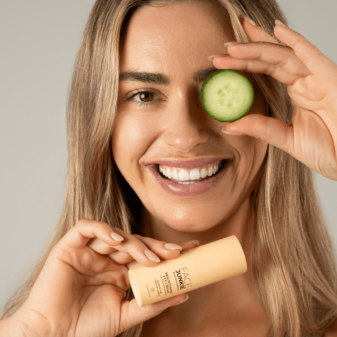 A smiling woman holds a cucumber slice over one eye while holding a bottle of &quot;FACE JUNKIE Brightening Eye Cream&quot; in her other hand. The woman has long, light brown hair and is showcasing a radiant complexion. The product is in a light peach-colored bottle, and the overall vibe of the image is fresh and playful, emphasizing skincare and eye care routines.