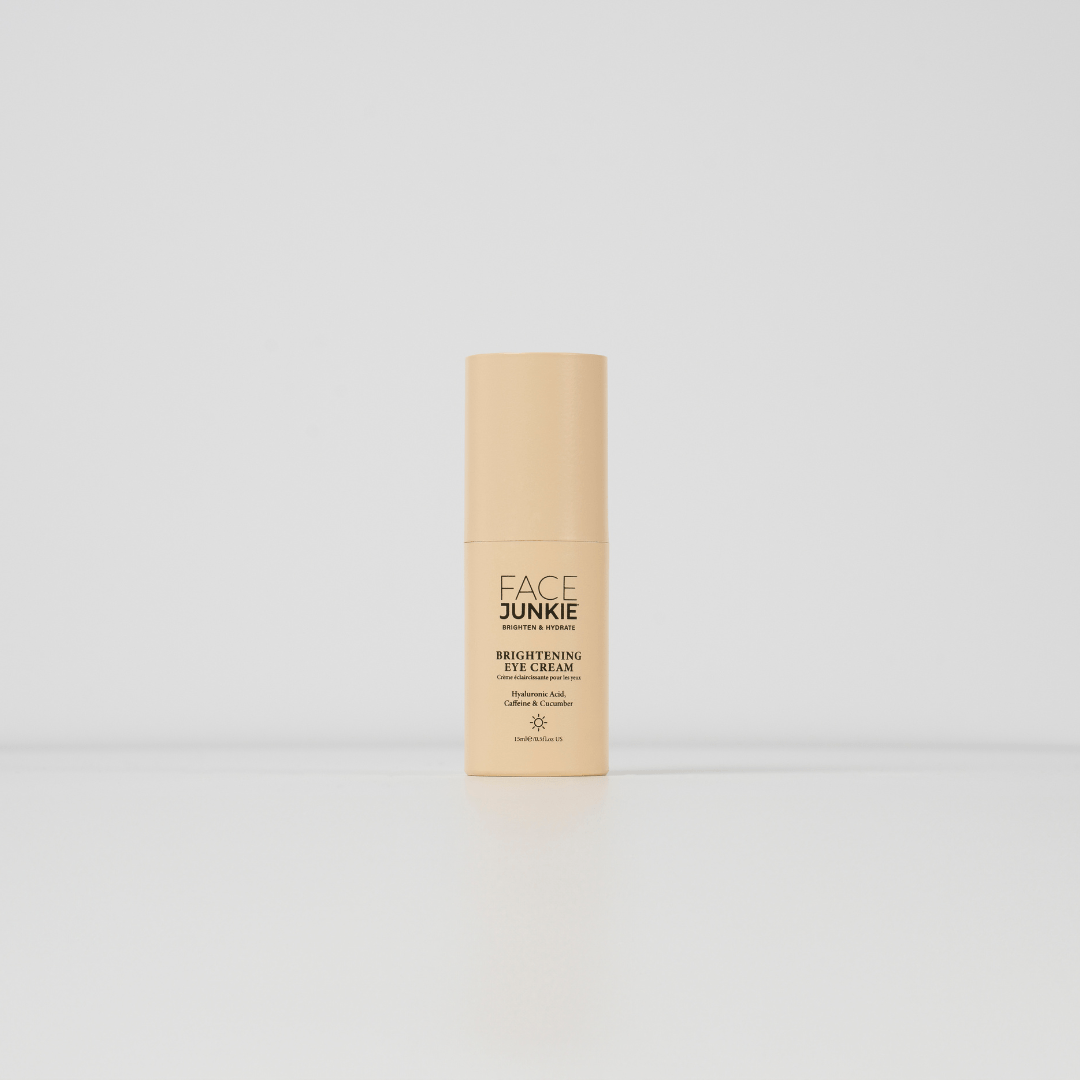 A minimalistic product shot of &quot;FACE JUNKIE Brightening Eye Cream&quot; in a light peach-colored bottle. The bottle stands on a clean white surface with a plain white background, emphasizing the product&