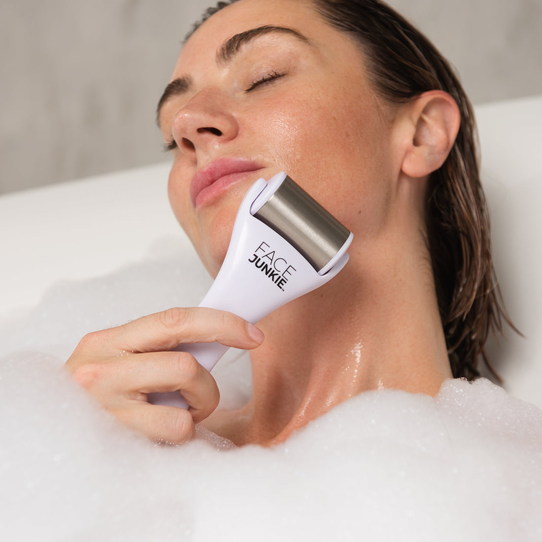 A woman relaxes in a bubble bath while using a Face Junkie-branded facial roller on her chin. Her eyes are closed, and her expression is calm, emphasizing the soothing, self-care aspect of the skincare routine. The stainless steel roller is gently applied to her skin, promoting circulation and a refreshing facial experience. The serene atmosphere and warm lighting highlight the rejuvenating benefits of incorporating the facial roller into a relaxation routine.