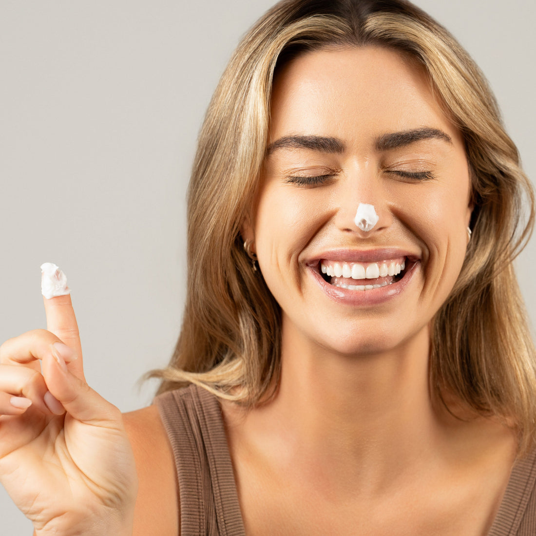 A smiling woman with light brown hair applies skincare cream to her nose, with a playful dab of the product on the tip of her nose. She holds up her finger, which also has a small amount of cream, while her eyes are closed in a joyful expression. She wears a brown sleeveless top, and the soft lighting highlights her glowing skin, conveying a sense of fun and happiness in her skincare routine.