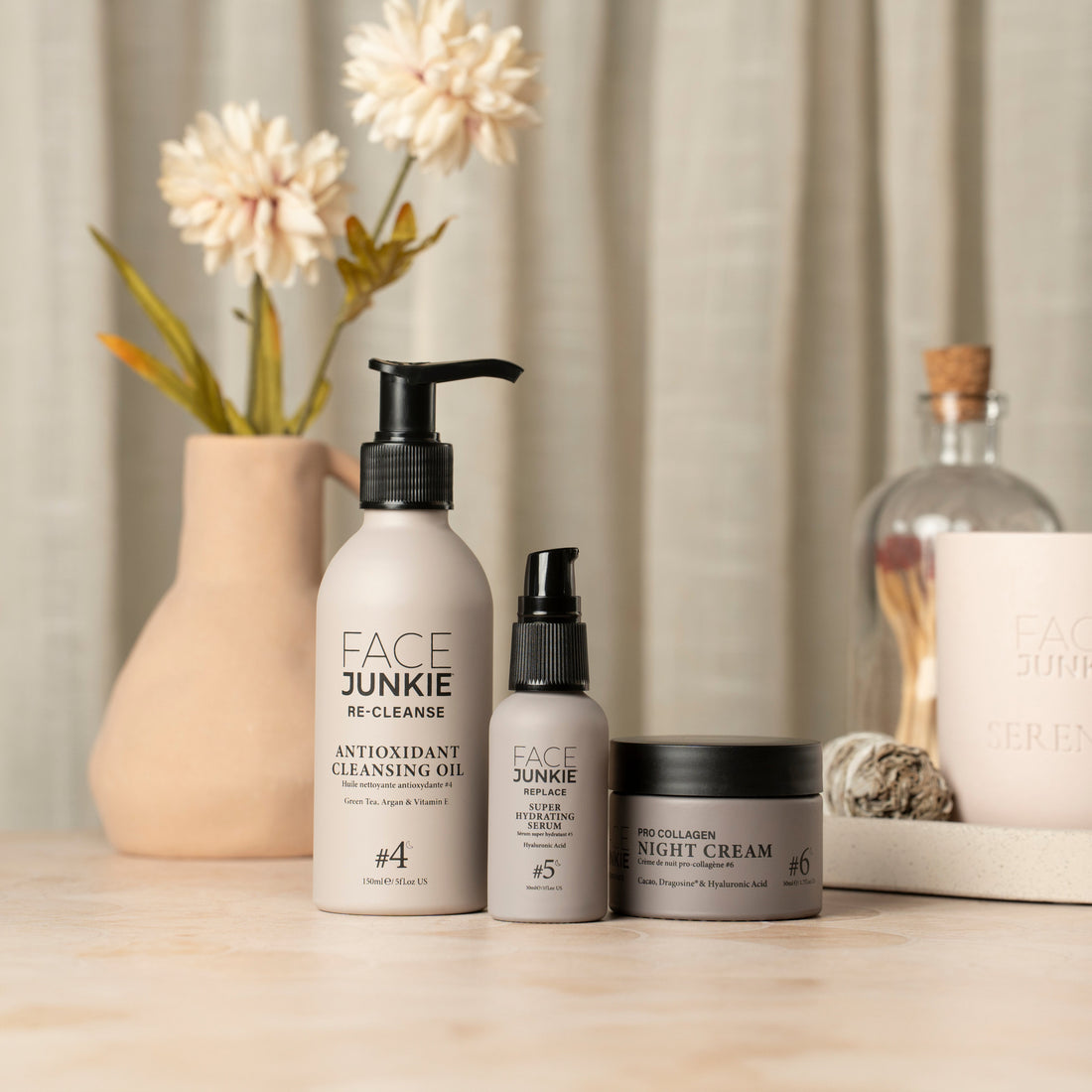 A trio of Face Junkie skincare products is elegantly arranged on a light wooden surface. The set includes Antioxidant Cleansing Oil, Super Hydrating Serum, and Pro Collagen Night Cream, labeled as steps 4, 5, and 6 in a skincare routine. In the background, a beige vase with soft white flowers and a decorative glass jar add a natural and calming touch, complementing the minimalist design of the products. The serene setting reflects a luxurious and peaceful skincare experience.