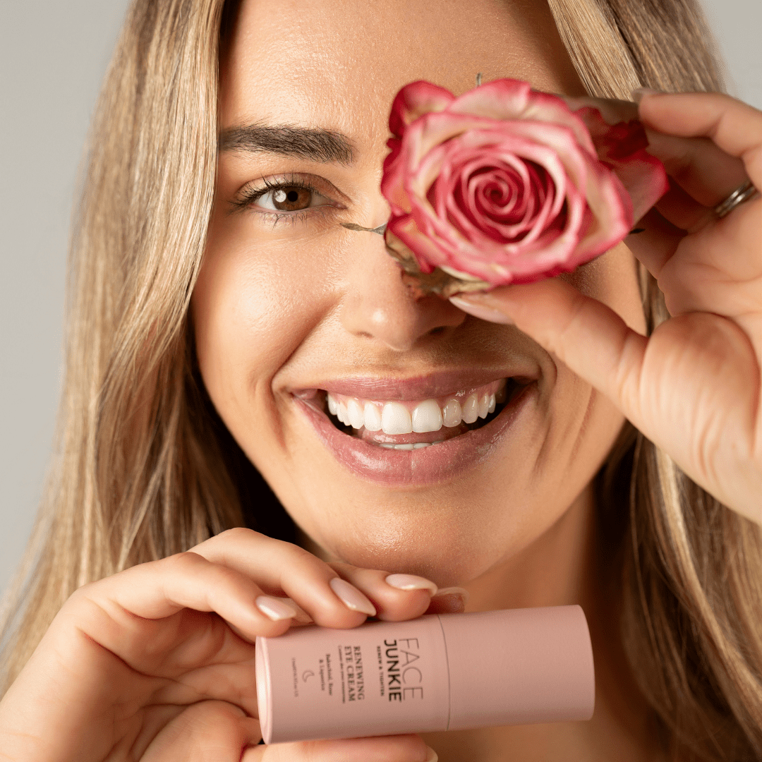 Picture of a blonde model smiling in an inviting manner while holding a rose to her eye and a renewing eye cream in her other hand