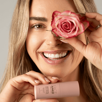 Picture of a blonde model smiling in an inviting manner while holding a rose to her eye and a renewing eye cream in her other hand