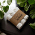 A set of rolled white towels wrapped in a Face Junkie-branded paper band is placed on a dark wooden surface. Surrounding the towels are lush green plants, creating a fresh, natural atmosphere. The soft lighting and clean, minimalist design emphasize the eco-friendly and luxurious feel of the towels, which are likely used as part of a skincare or self-care routine. The contrast between the white towels and the green leaves adds to the serene and refreshing ambiance.