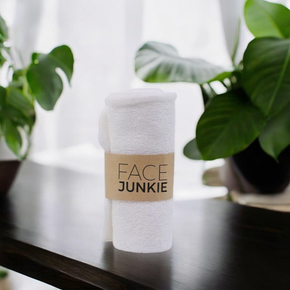 A rolled white towel with a Face Junkie-branded paper band around it is placed on a dark wooden surface. The background features lush green indoor plants, creating a fresh and serene atmosphere. Soft natural light filters through sheer curtains, enhancing the clean and minimalist aesthetic of the scene. The towel suggests a luxurious and eco-conscious addition to a skincare routine.