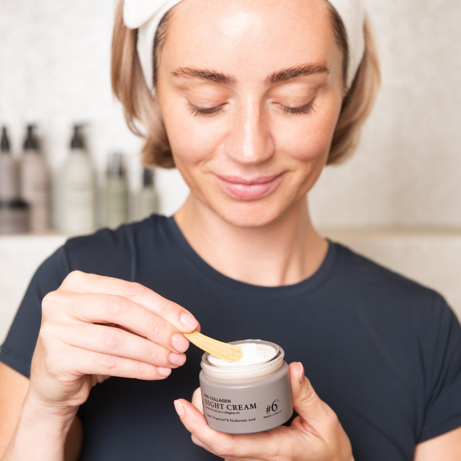 Jen Graham, known as Charity Shop Girl, is smiling gently as she scoops a small amount of FACE JUNKIE night cream from a jar using a wooden spatula. She wears a white headband and a dark top, standing in a bathroom with a row of skincare products blurred in the background. Her expression is calm and focused, showcasing a self-care moment as she prepares to apply the product. The night cream jar is labeled &quot;