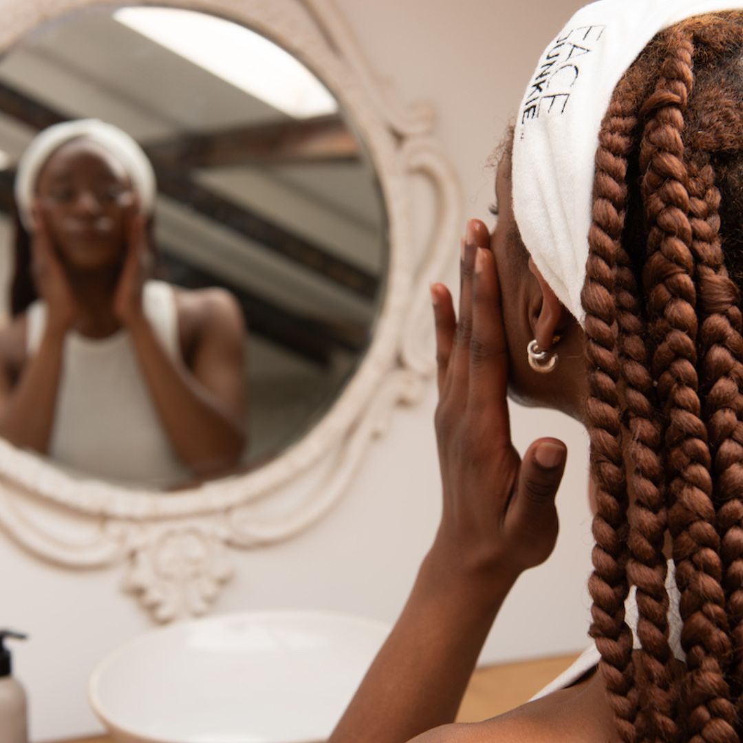 A woman with long, braided hair is seen from behind as she gently applies skincare while looking into an ornate mirror. She wears a white Face Junkie-branded headband, keeping her hair back as part of her skincare routine. The scene takes place in a well-lit bathroom, with a sink and skincare products visible on the counter. The reflection in the mirror shows her carefully following her skincare regimen, reinforcing a sense of self-care and focus on healthy skin.