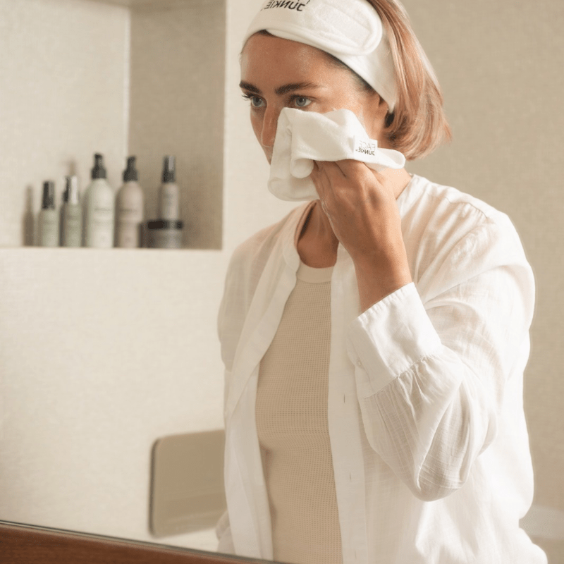 A woman stands in front of a bathroom mirror, gently wiping her face with a FACE JUNKIE cleansing cloth. She wears a white headband with the brand&